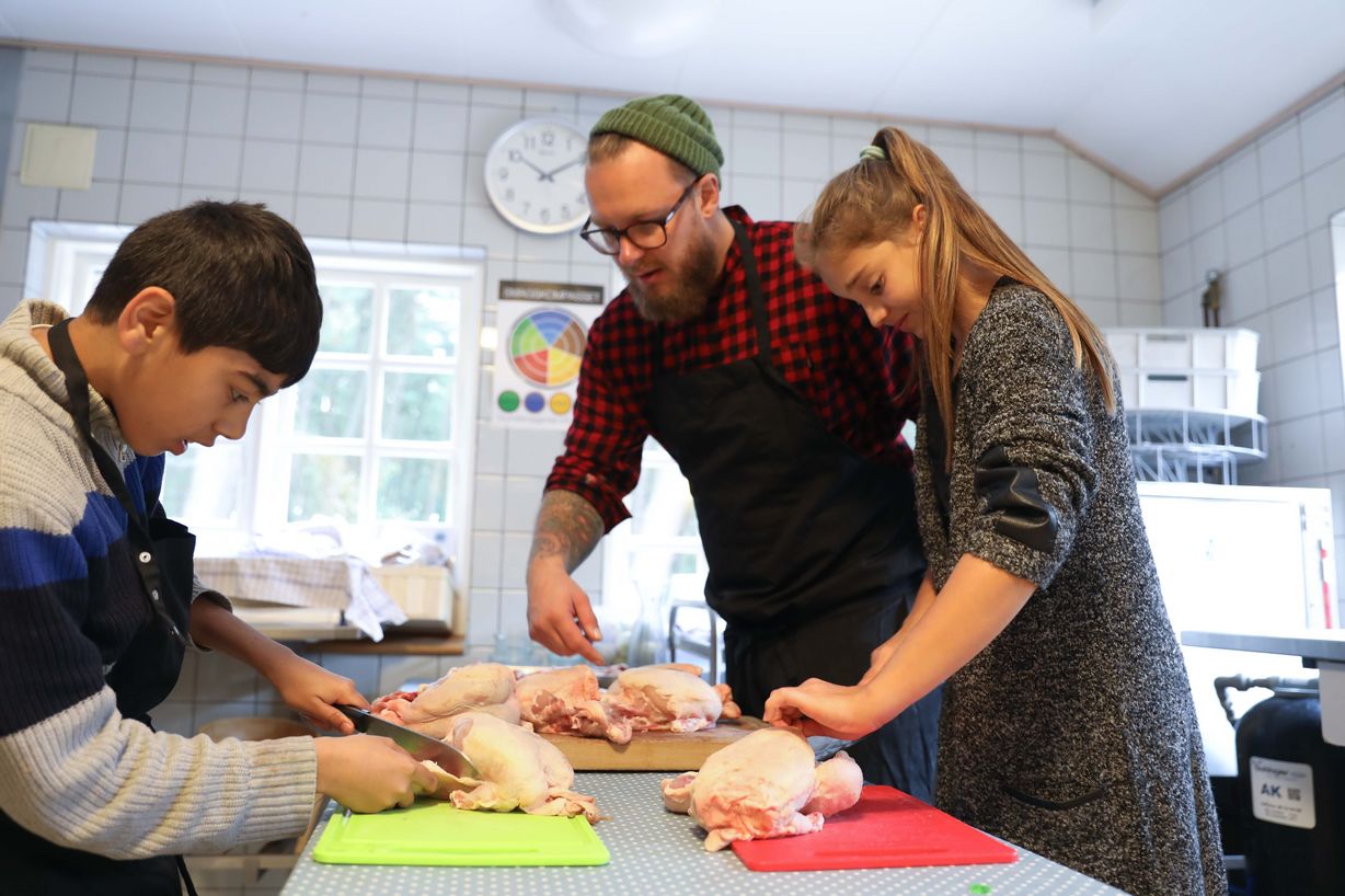 Anees (tv.) er sammen med Milica (th.) i køkkenet for at forberede aftenens festmenu. Kokken Martin 'Guf' Becker vejleder eleverne i korrekt udskæring af kyllingerne. Foto: Jan Klint Poulsen